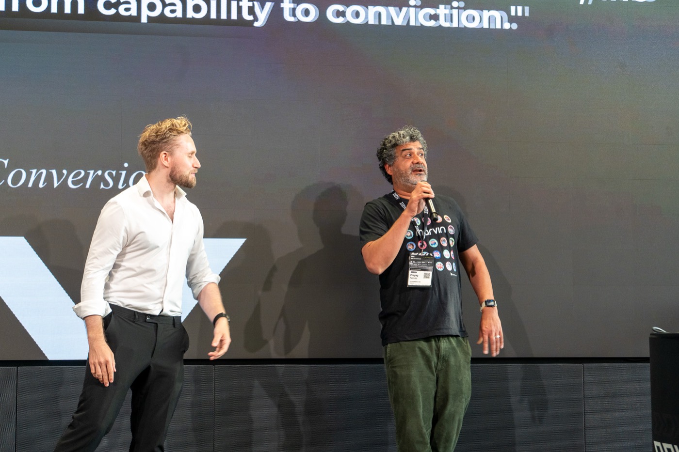 Two speakers on stage under a screen reading from capability to conviction